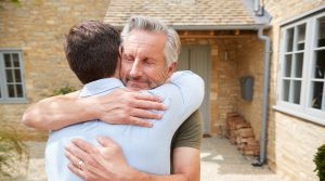 Senior father greets and hugs adult son outside front door of house as he visits