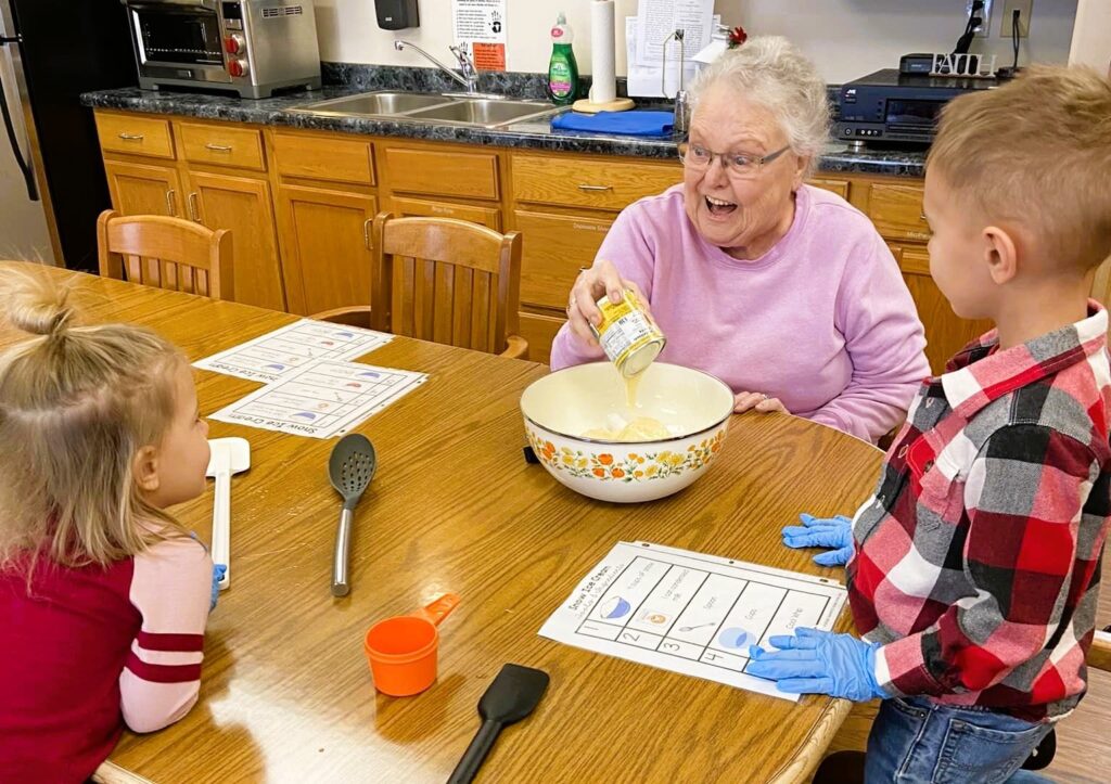 A senior woman adding ingredients and smiling as the kids watch her with fascination.