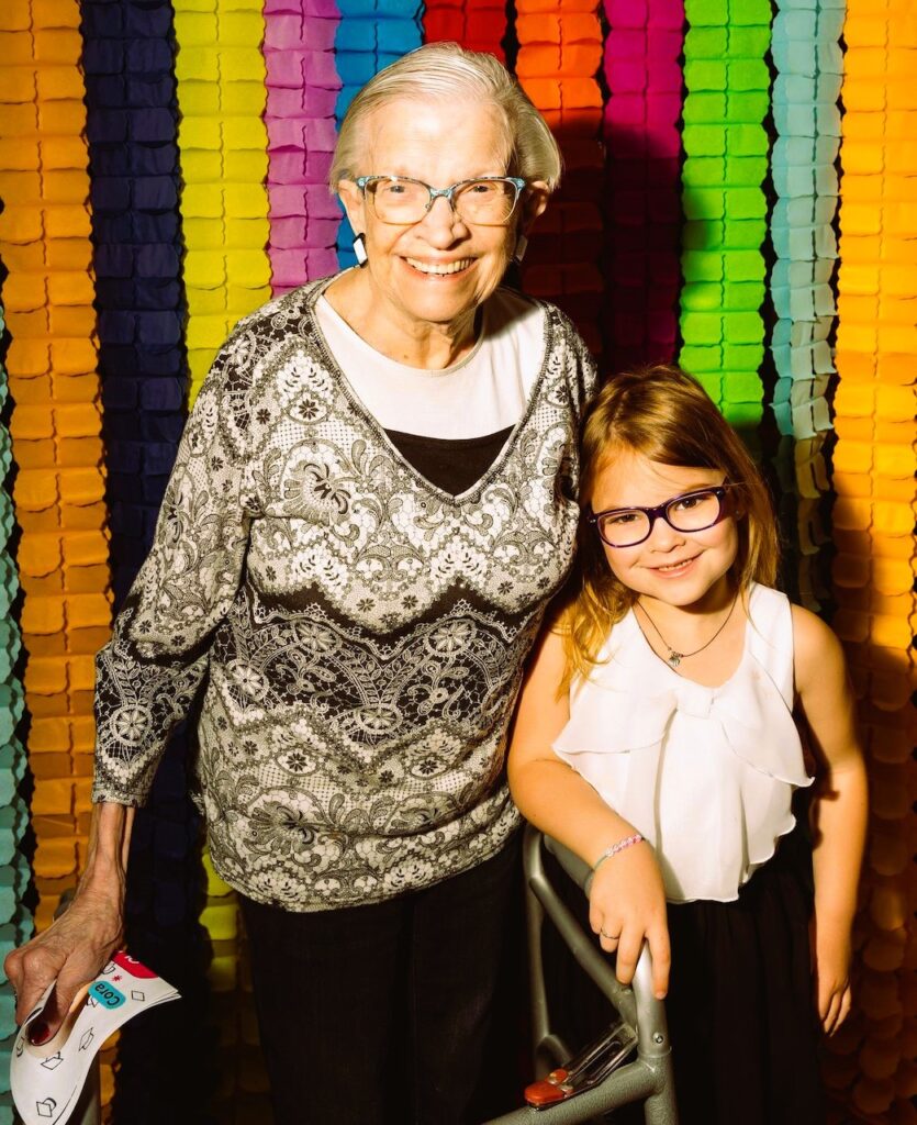 A senior woman and little girl smile and pose together in front of a colorful wall